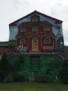 Mural on a building depicting an old-style train depot, with a couple of waiting passengers, a dog, and an arriving steam train.