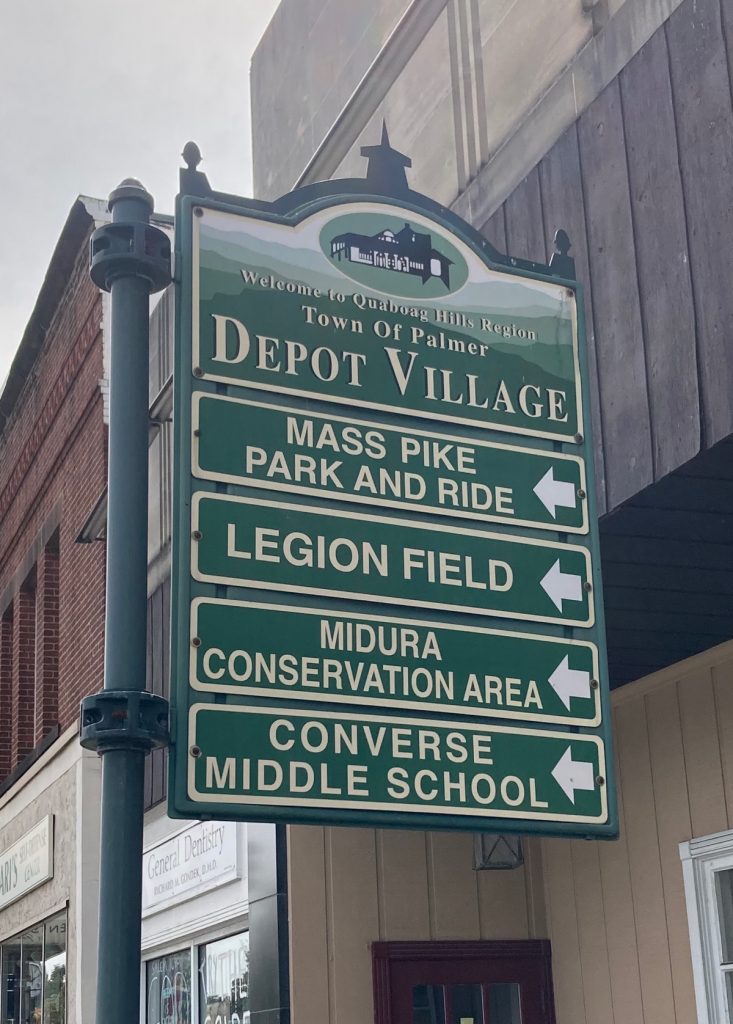 Green sign in front of downtown buildings.  The sign reads "Welcome to Quaboag Hills Region - Town of Palmer Depot Village", and below that has arrows directing viewers to various places in town.