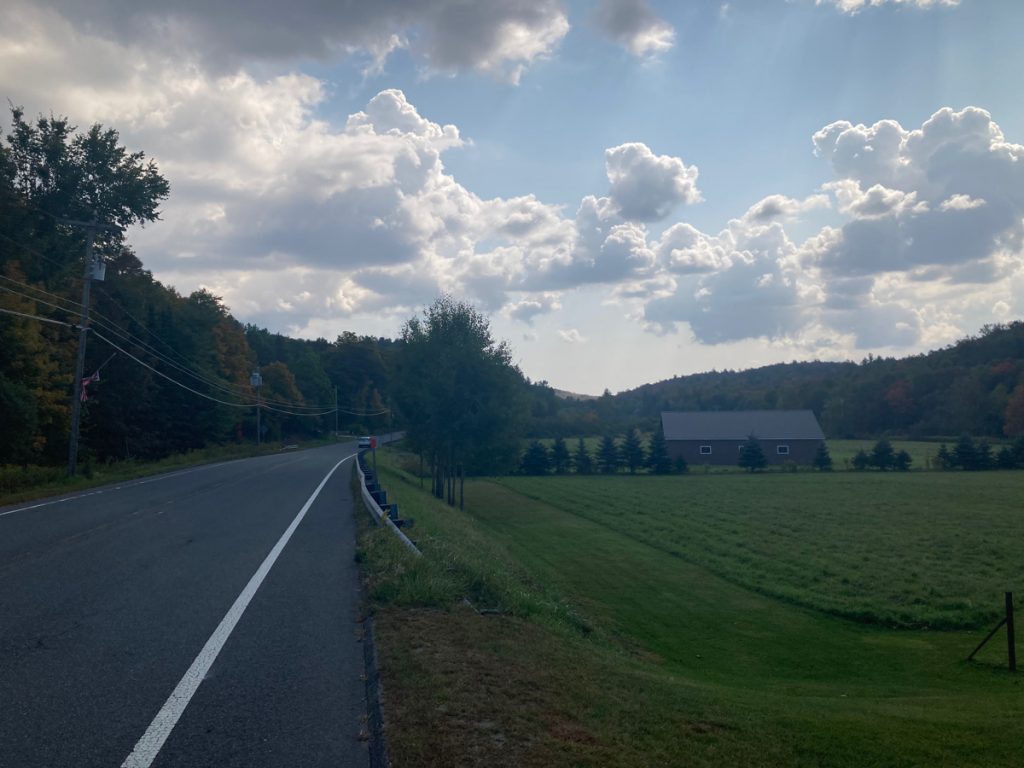 Road surface on the left, with a green field and barn to the right.  There are hills and trees in the distance, and a bunch of clouds in the sky.