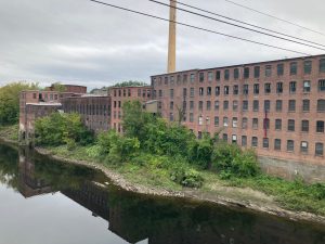 Old brick mill buildings stretch along the bank of a river, with various bushes and brush growing between the water and buildings.  There is a single tall smokestack in the center of the photo.