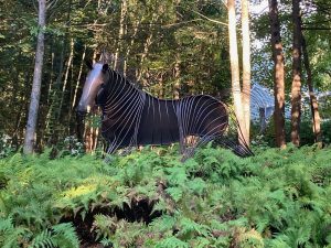 A sheet metal and wire sculpture of a horse, with many ferns growing in front of it, and trees behind.