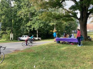 Students and cyclists sit and stand around a pair of tables with purple tablecloths.  The tables are under a tree on a grassy area next to a driveway,and there are other trees behind. 