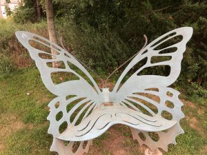 Frontal view of a bench shaped like a butterfly, in front of some bushes.