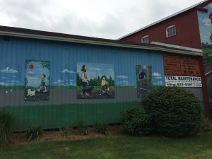 Mural on the side of a building, depicting different types of trail users, including a child walking with a dog and balloon, an adult walking with a dog, a child on a bike with training wheels, and a skateboarder.