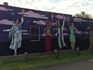Mural on the side of a building, depicting five women of different ages, races, and ethnicities, each holding a burning torch aloft in one hand.