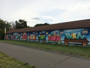 Mural depicting eight joined train cars, with different items in each car.  The last one is carrying large letters spelling the name Ben.
