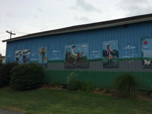 Mural on the side of a building, depicting different types of trail users (3 cyclists, a jogger, and a rollerblader).  There is some landscaping at the base of the wall.