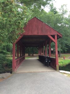 Red, wooden, covered bridge over a small stream, with trees and picnic areas on the other side.