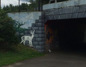 Mural of a deer and small cityscape, as well as painted stones on the concrete wall of a bike tunnel entrance.