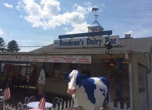 Building labeled "Rondeau's Dairy Bar", with a blue and white cow statue in front of it.