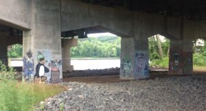 Graffiti of a penguin with a fire in a bareel, and a green caricature head, painted on concrete supports of freeway overpass.