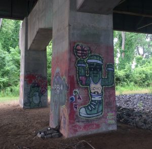 Graffiti of a cactus dressed as a basketball player, with basketball,  and a dinosaur with a big foam #1 hand, painted on concrete supports of freeway overpass.