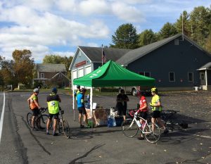 Food and water table under a canopy, with half a dozen cyclists around it.  This is in a parking lot, with the Guilford fire station in the background.