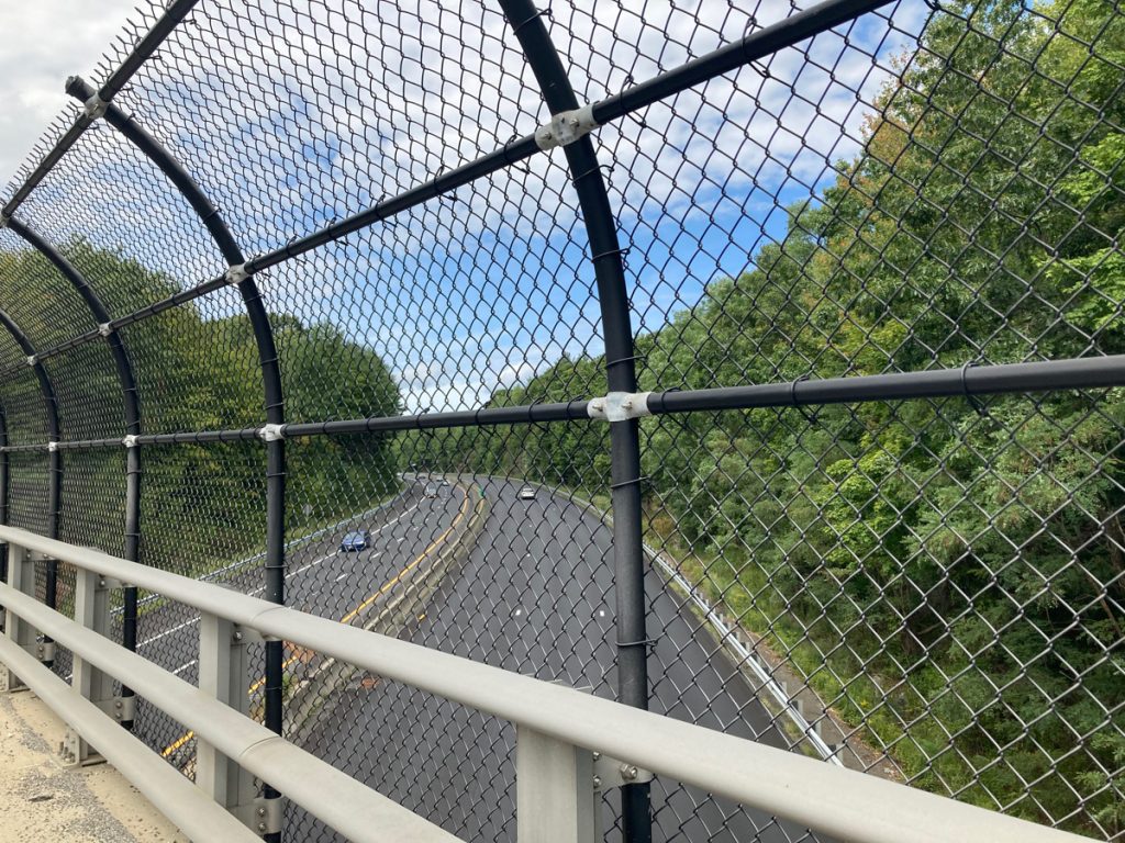 Freeway below, seen from an overpass, looking through chain-link fence.  Trees line the expressway.