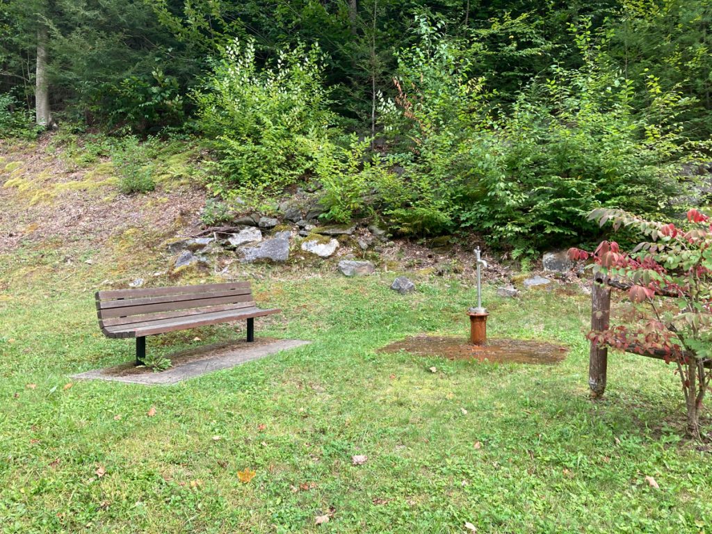 Wooden bench near a water spigot on a rusty base, in a grassy area with bushes and trees behind. 