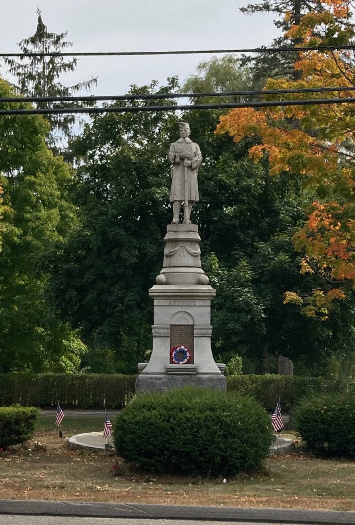 Stone monument with a statue of a man at the top.  Lettering on the base reads "Argonne".  There are bushes, trees, and grass around the monument.