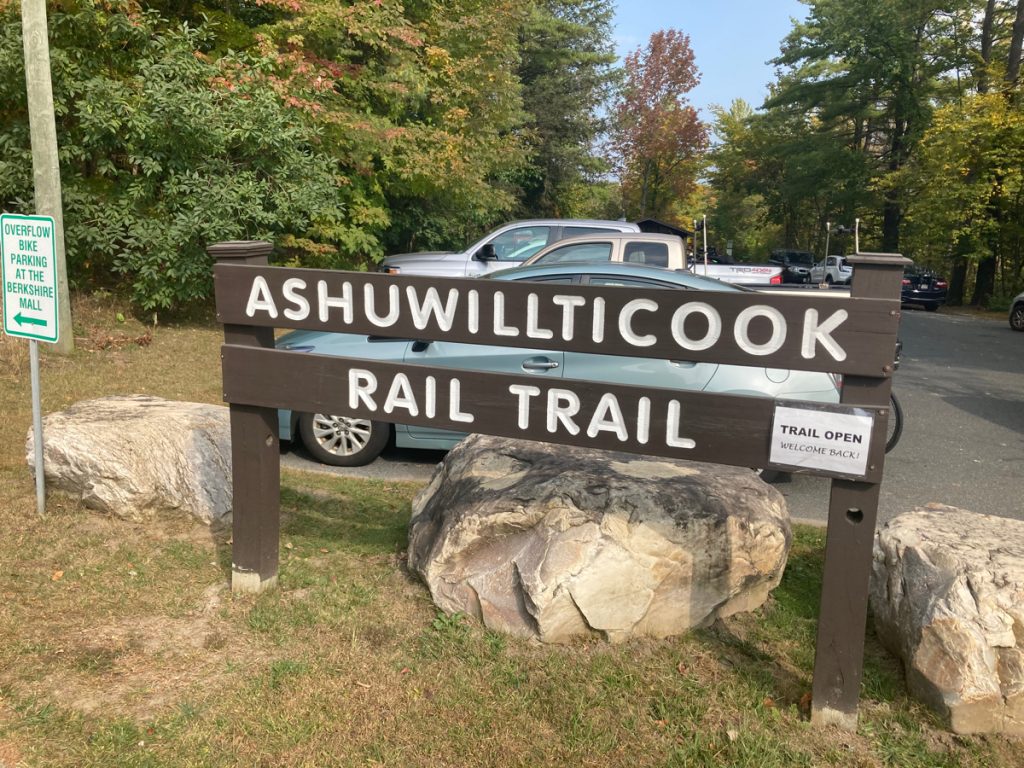 Wooden sign with white lettering reading "Ashuwillticook Rail Trail" with large rocks around the sign posts.  There are parked cars and trees behind it.  