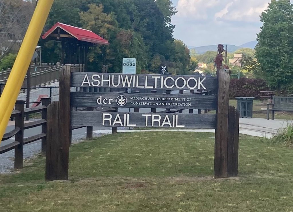 Wood sign with carved, white-painted letters reading "Ashuwillticook Rail Trail - Massachusetts Department of Conservation and Recreation".  Around the sign are grass and trees, and some paved trail surface with a wooden fence.
