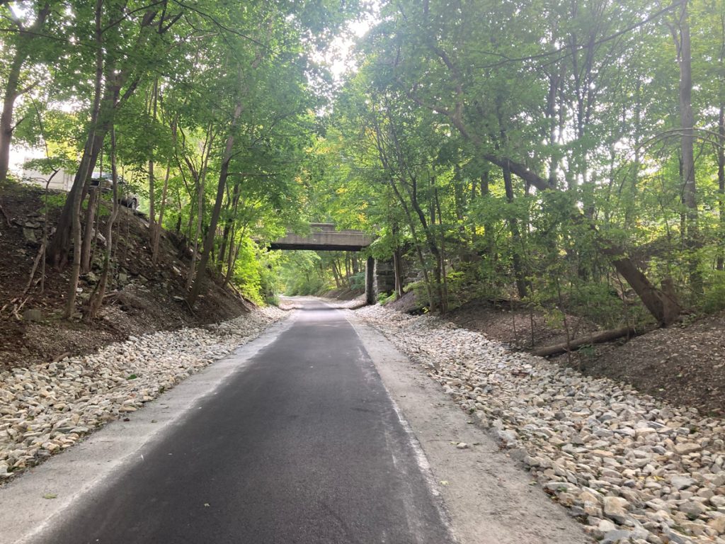 Looking along paved trail with rock beds on each side, then further flanked by trees.  Up ahead there is a bridge over the trail for a road.