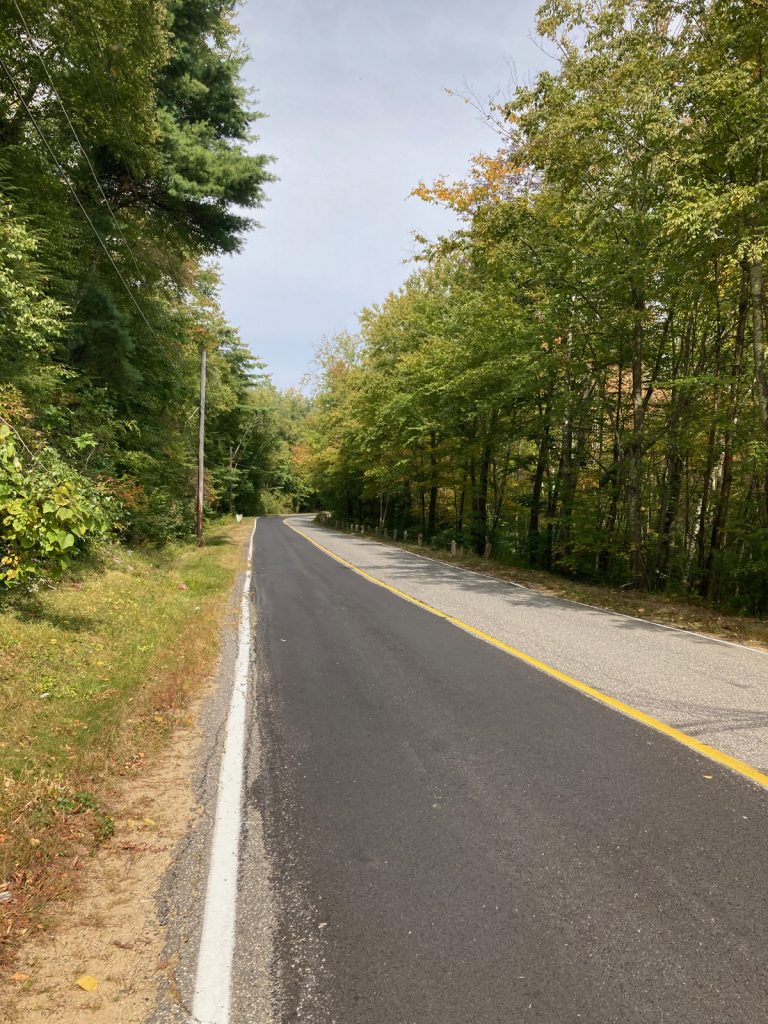 Stretch of road with one lane of dark blacktop, and the other lane lighter in color.  There are trees on each side of the road.