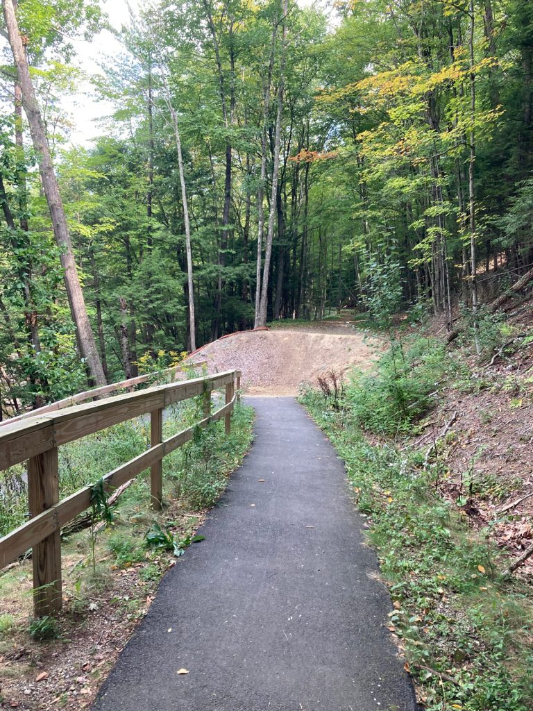 Narrow paved trail stretching a short distance away, with a wooden railing on the left side.  Ahead is a dirt berm, and trees beyond that.