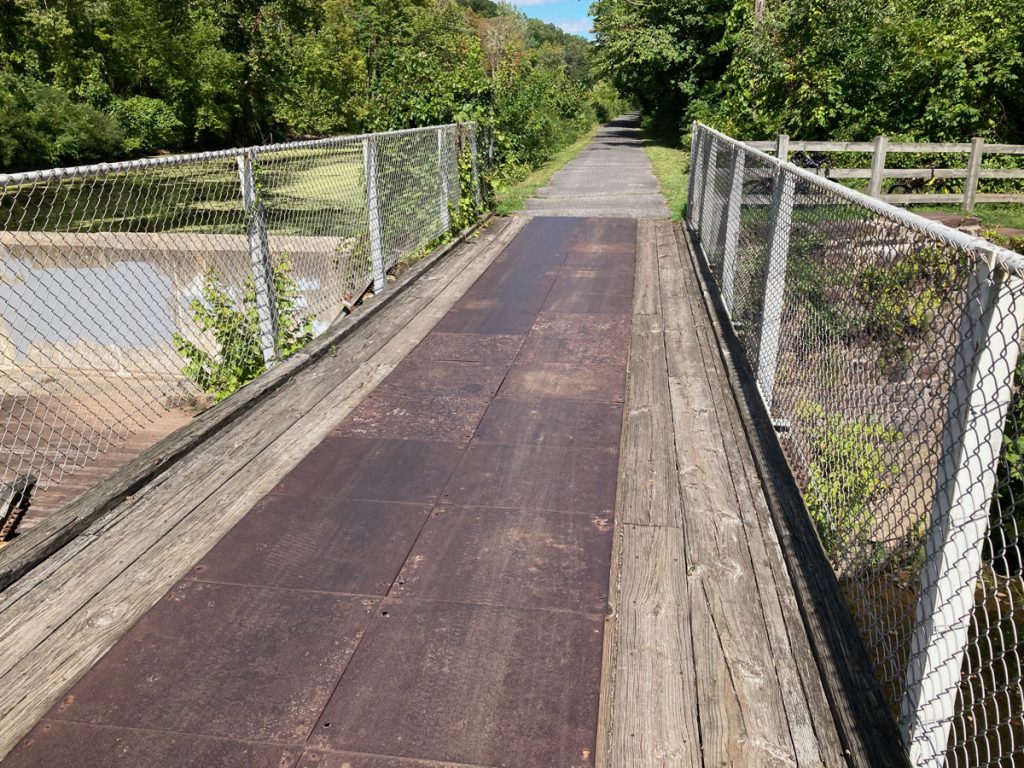 Looking along bike trail bridge with wood and metal deck, and chain link fence on either side.