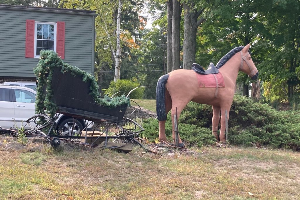 Horse figure on a lawn in front of a parking lot, with an ivy-covered cart or sleigh hitched up to it.  A part of a building is in the background.