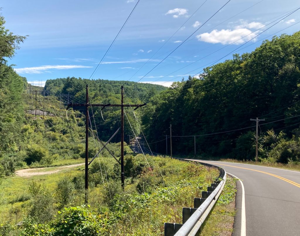 Looking downhill with a road on the right, grass and brush on the left, and some electric wire towers stretching into the distance, heading over a tree-covered hill (through a gap in the trees on the hill).