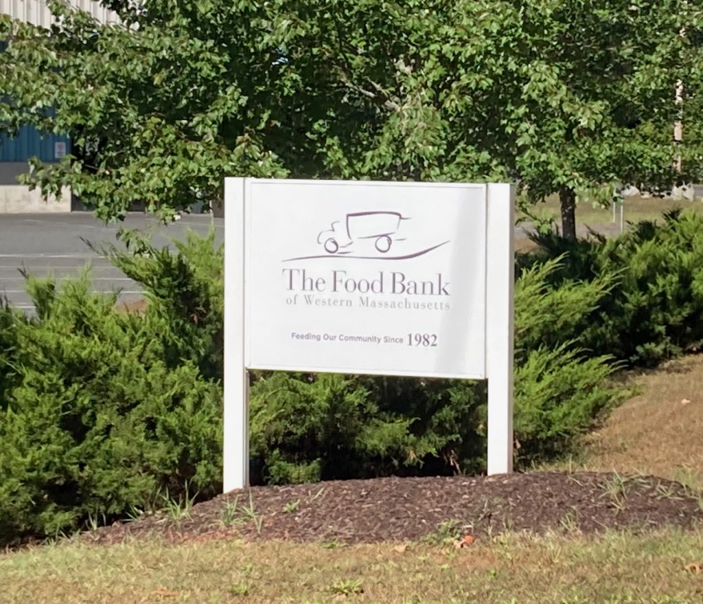 White wooden sign with text reading "The Food Bank of Western Massachusetts" and "Feeding our community since 1982", as well as the Food Bank's logo.  The sign has shrubs and trees behind it, as well as a bit of parking lot.