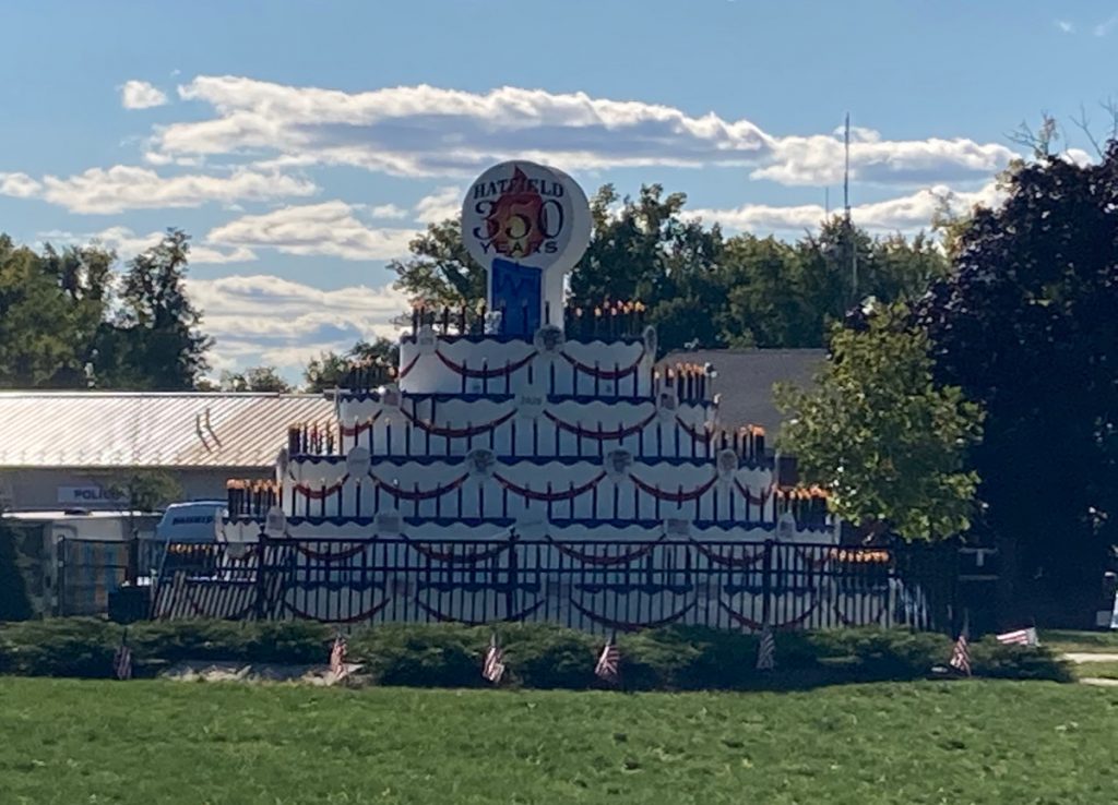 Large outdoor sculpture of a birthday cake, decorated in red, white, and blue, with a sign at the top reading "Hatfield 350 years".