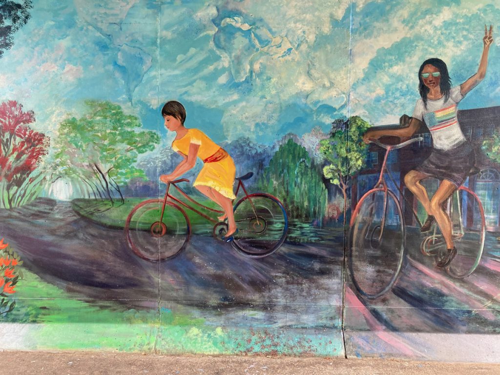 Mural panel depicting cyclists on a recreation trail, one of them waving to the viewer
