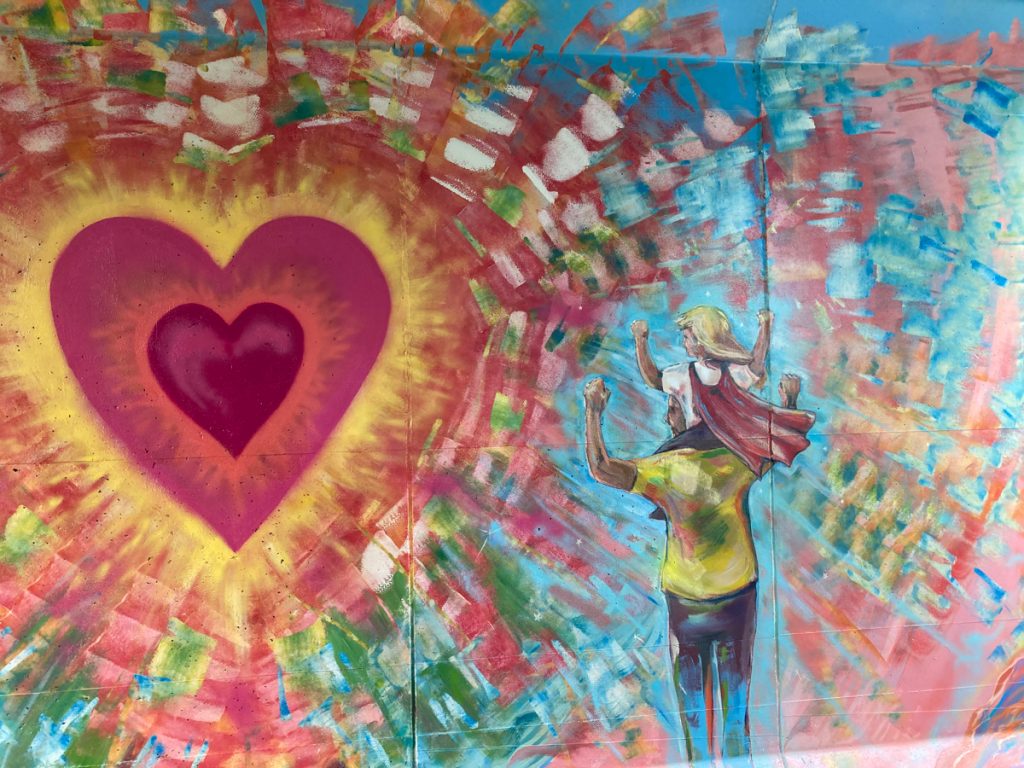 Mural depicting large red heart that is radiating color, with an adult facing it, and a child on the adult's shoulders.  Their hands are raised in the air.