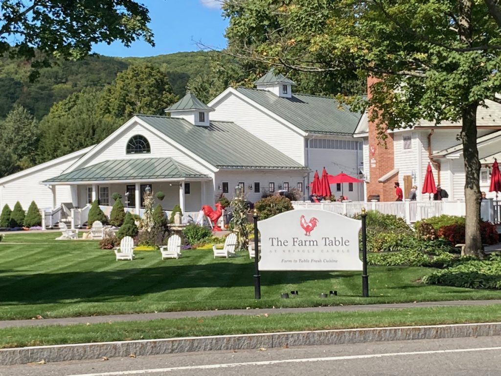 White building with flowers and other landscaping in front of it, as well as a red rooster statue.  A sign in the foreground (by the roadside) reads "The Farm Table at Kringle Candle", and has a red rooster logo.