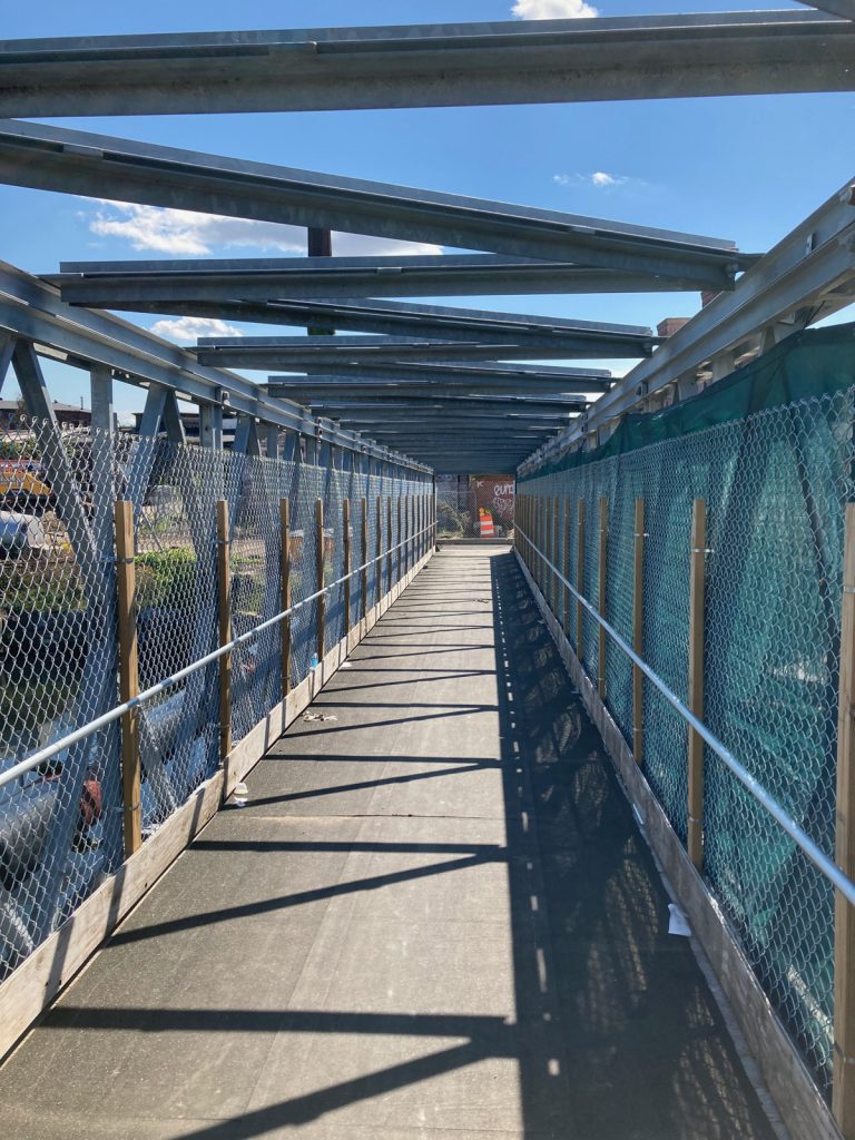 View along pedestrian bridge, there are metal beams and fencing on either side, and beams crossing the top of the bridge structure