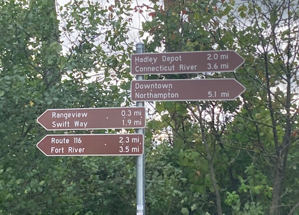 4 brown signs on a post, with white text indicating the distance and direction to several places, such as the Connecticut River, downtown Northampton, and the Fort River.