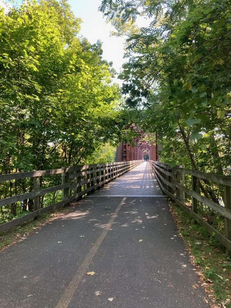 Looking along paved trail toward rusty-frame metal bridge (with wooden deck), and trees on either side of the trail.