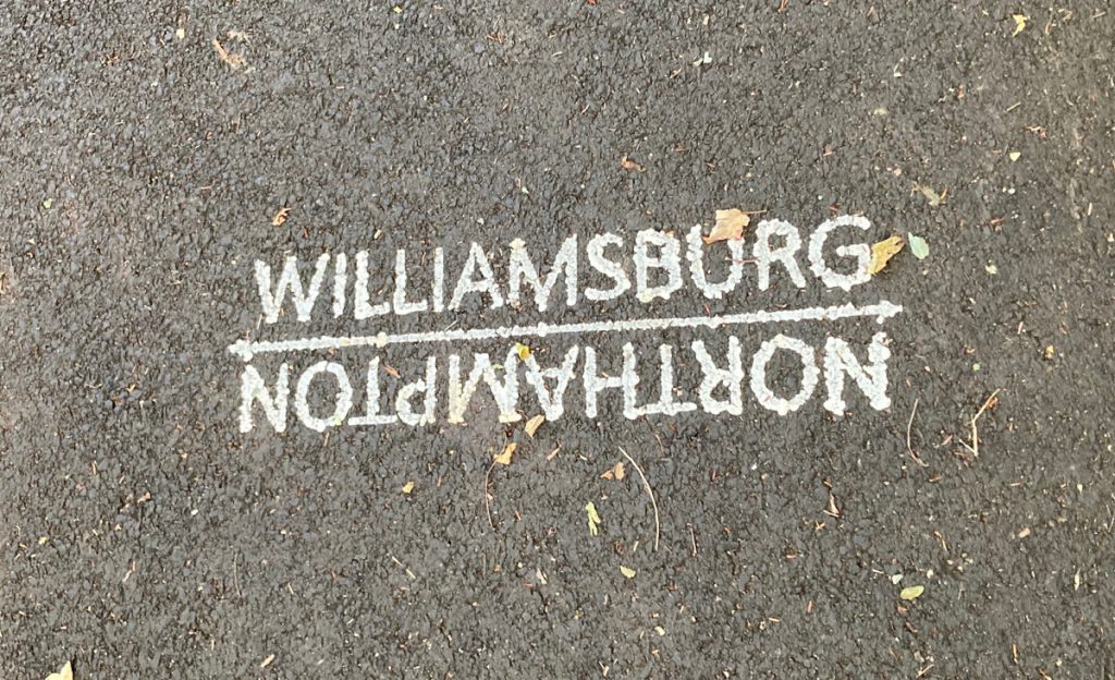 White paint on pavement which reads "Williamsburg" on one side of a line, and then upside-down lettering on the other side of the line, reading "Northampton".