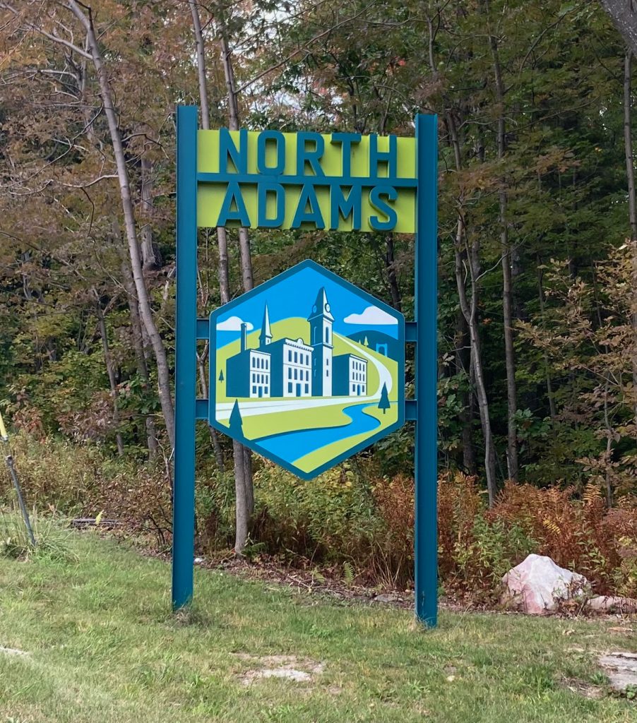 Tall blue and green sign reading "North Adams", with a stylized image of old industrial buildings by a river and hills.  The sign stands in some grass by the edge of a wooded area.