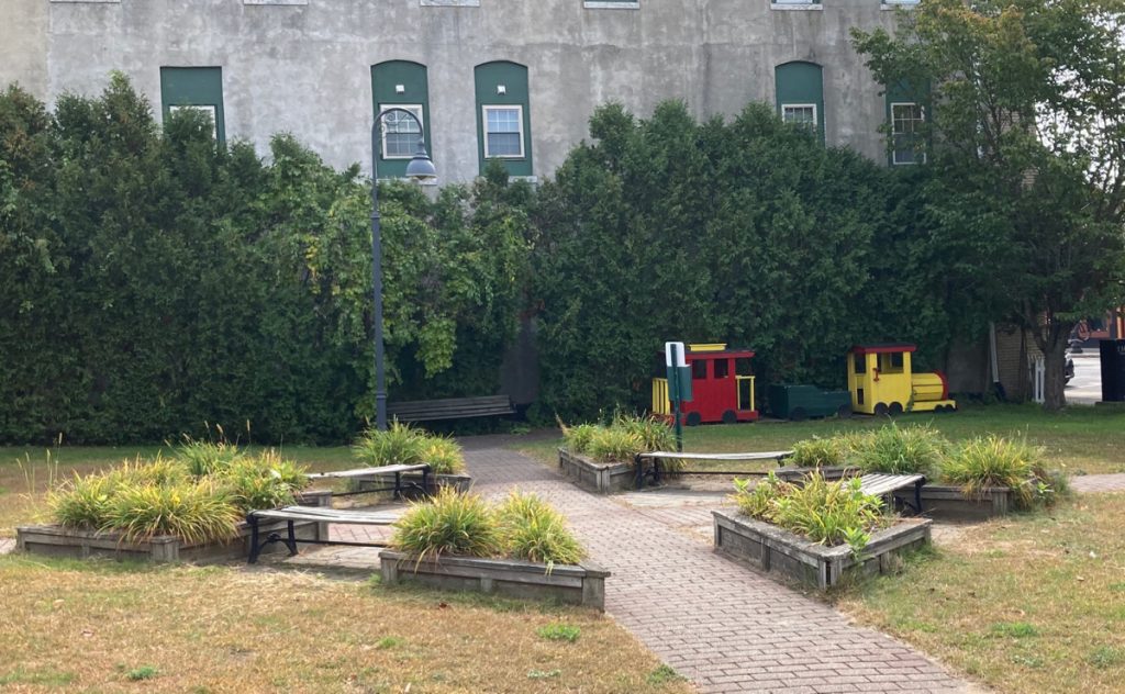 Small park with grass, brick walkway, benches, raised planters with perennials, and a small red and yellow play train.  Behind all of that are some small trees and a 2-story building.