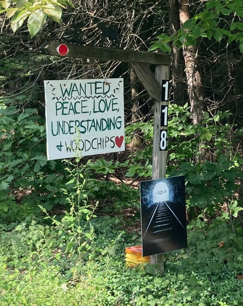 Sign hanging from a wooden post that reads "Wanted: peace, love, understanding, and wood chips".  There is greenery all around.