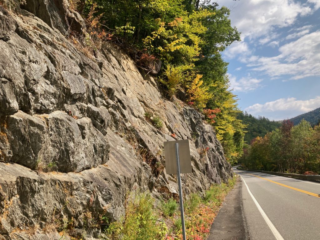Rock face to the left of a road, with trees at the top of the rocks.  Some trees can be seen on the right side of the road.