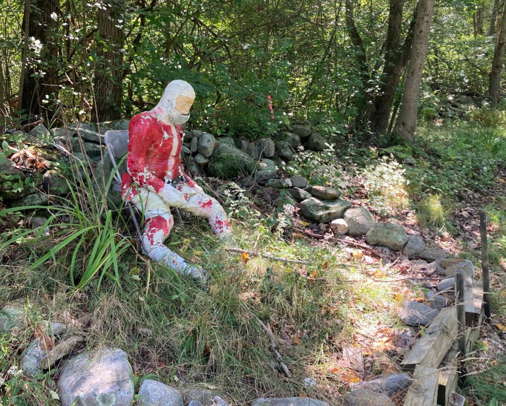 A very weathered, stuffed figure looking like Santa Claus, wearing a medical face mask, seated in grass and surrounded by a ring of stones.