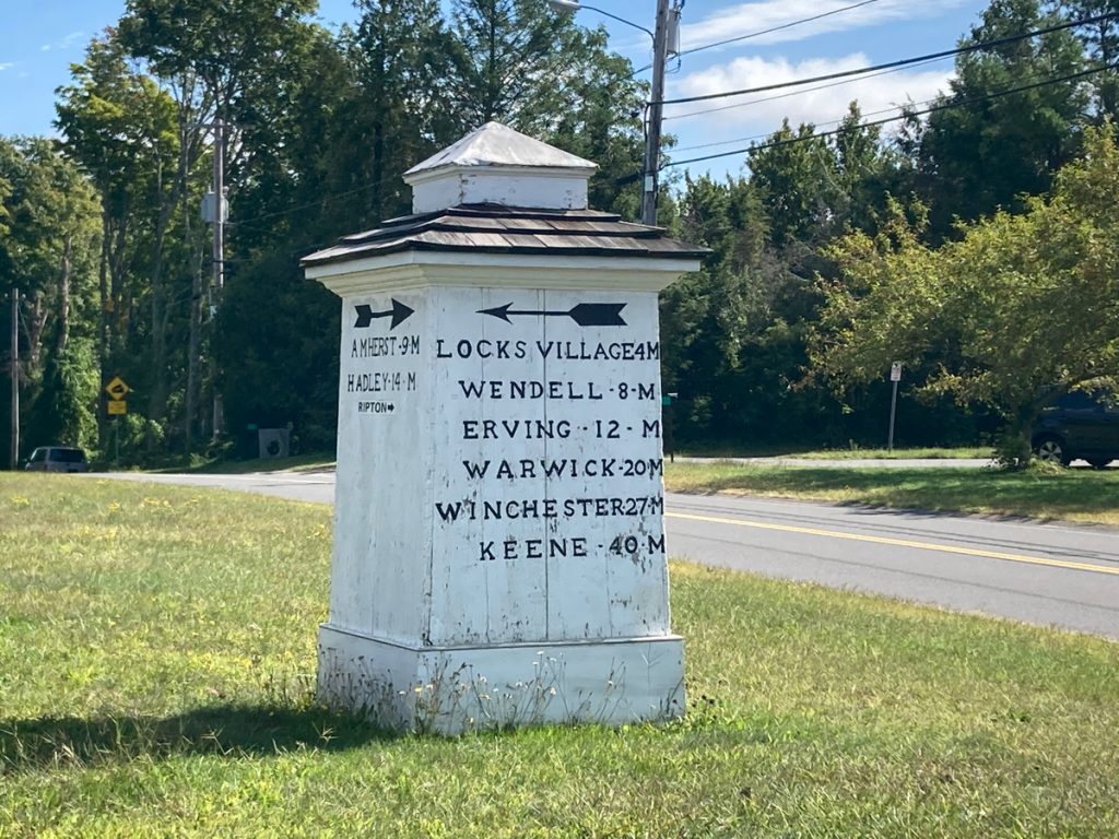 White, wooden 4-sided sign column, giving distances to different towns and areas in black text.  It stands in a field of grass and a road passes nearby, with trees in distance.