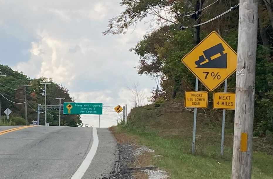 Road surface on the left with a yellow sign on the left warning of a 7 percent grade for the next 4 miles, and advising trucks to use lower gear.  In the distance there is a green sign above the road reading "Steep Hill - Curves Next Mile - Use Caution".