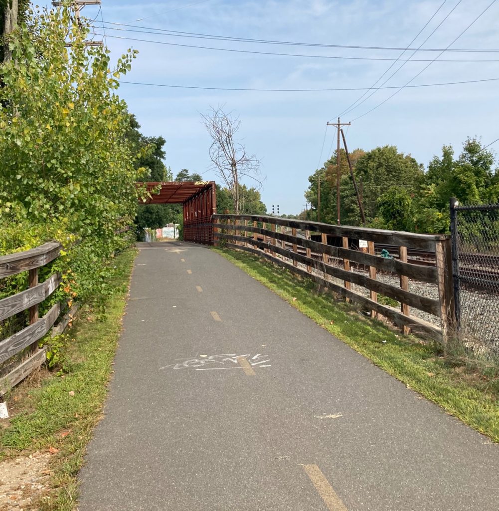 Looking along paved trail, there are wood fences on either side and the rusty-red frame of a bridge in the distance.  Railroad tracks can be seen to the right.