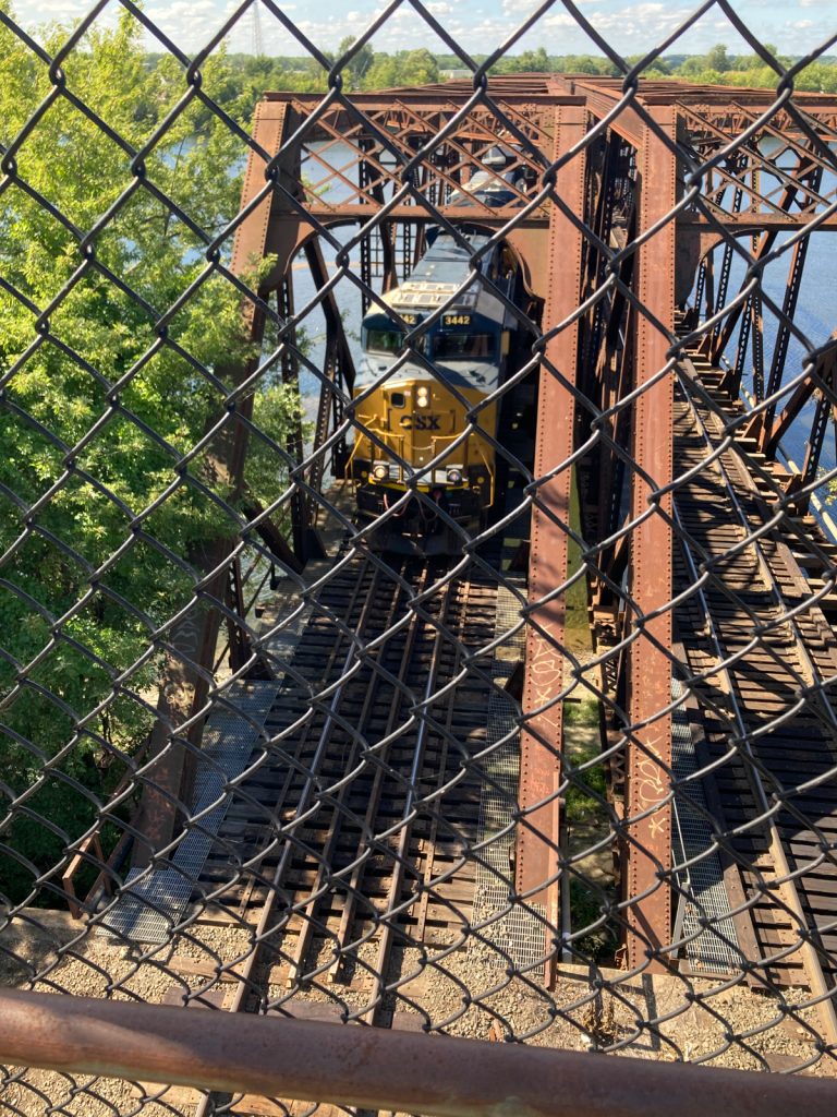 Looking down through chainlink fence at a train coming off of a bridge, toward the camera