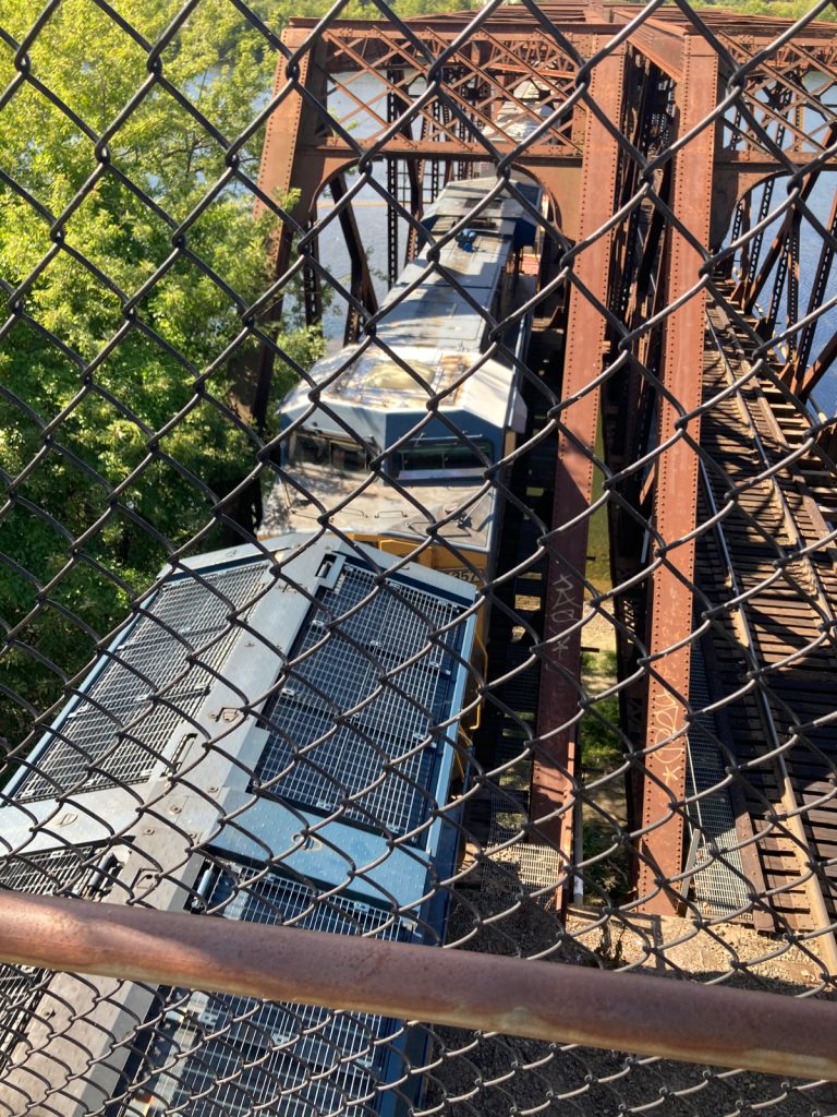 Looking down through chainlink fence at a train coming off of a bridge, passing under the camera