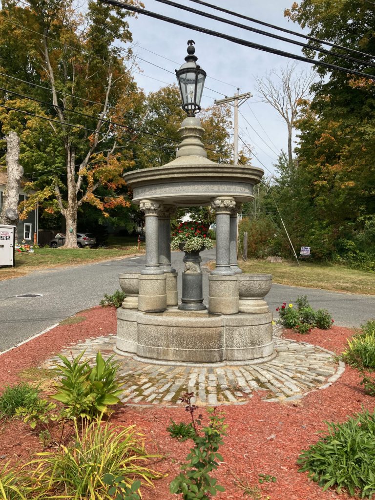 Stone monument which includes four pillars on a round base, with a planter containing flowers between the columns, a platform on top of the columns, and that is topped off by a glass and iron lamp.  There is some landscaping with red wood chips around the monument, with a road and trees in the background.