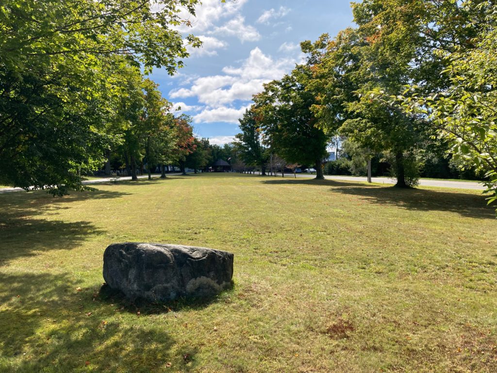 Long field of grass on town common.  There is a large rock in the foreground, and trees line each side of the common.