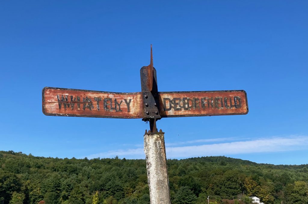 Rusty metal sign reading "Whately" on the left and "Deerfield" on the right.  Blue sjy and trees on hills are visible behind.